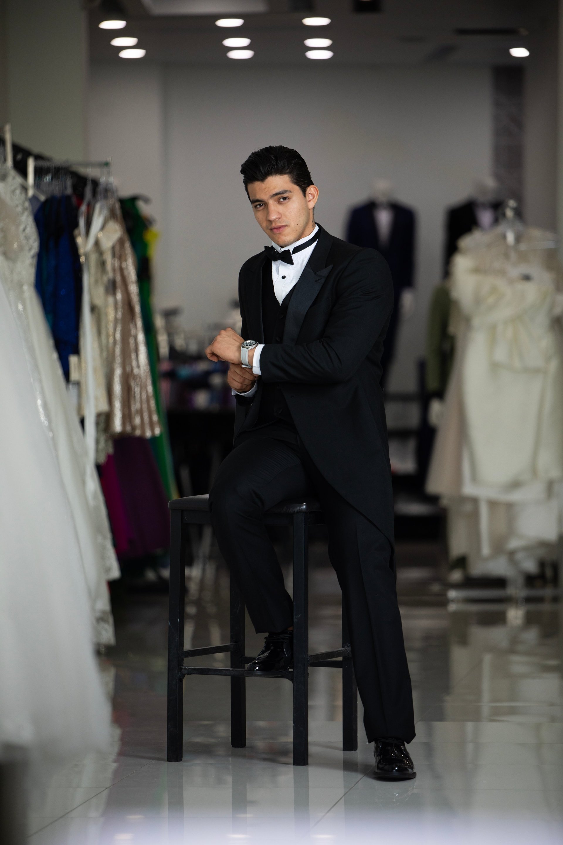 Man in black tuxedo and bow tie sitting on stool in fashion showroom surrounded by clothing displays