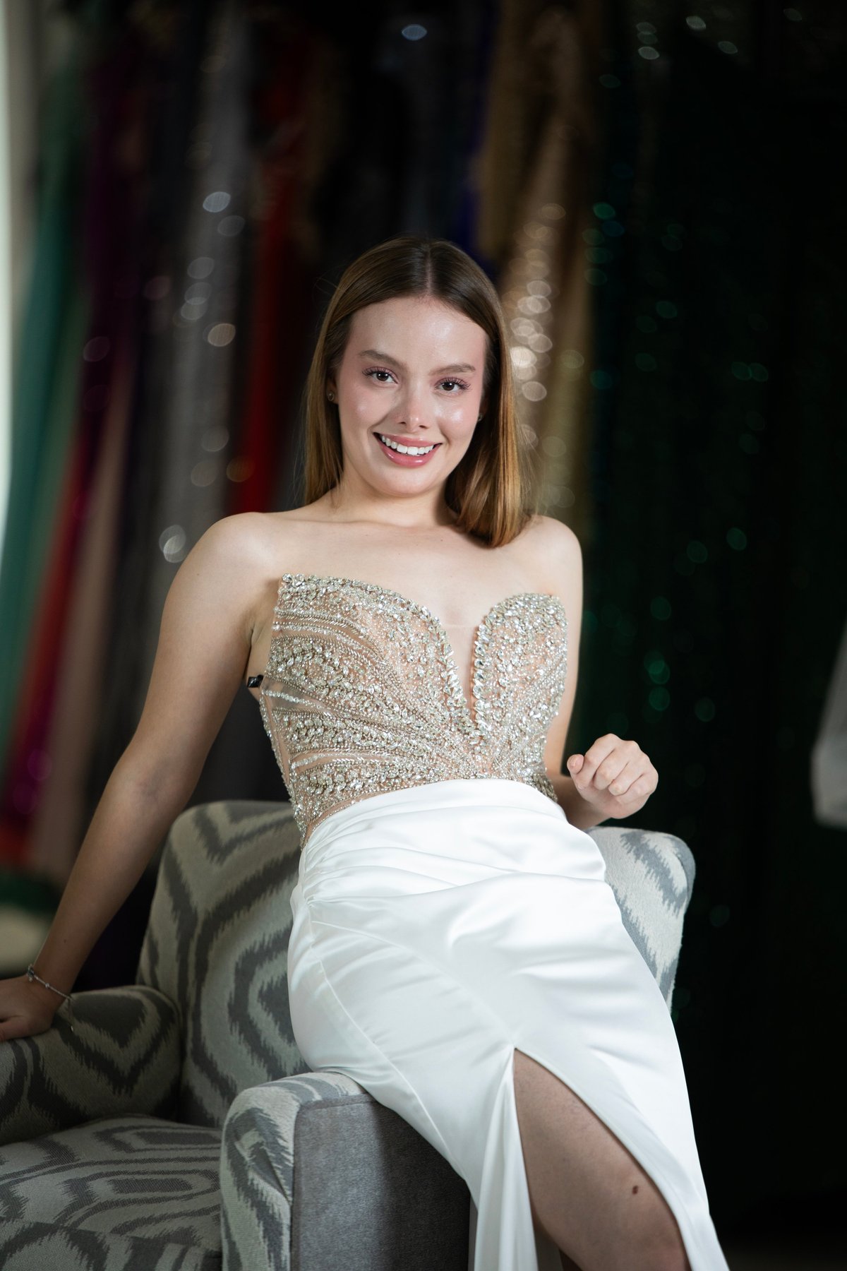 Mujer joven con cuerpo de vestido champagne con beads elegante con falda blanca sonriendo mientras posa en silla contra fondo oscuro con telas coloridas