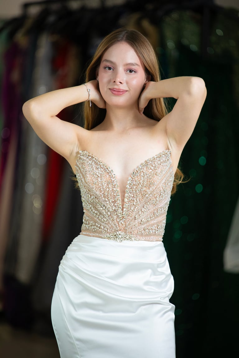 Mujer joven con top dorado con beads y falda blanca posando con manos detrás de la cabeza frente a percheros