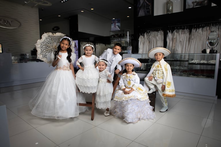 Group of children wearing elaborate white communion or formal dress costumes with large white hats and feathered accessories in an elegant room setting