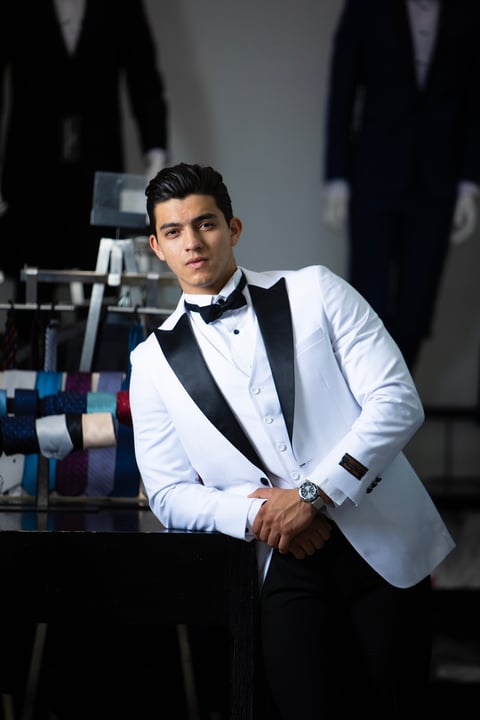 Man in white tuxedo jacket with black bow tie and dress pants leaning against glass surface